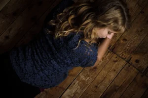 Top-down view of a woman with wavy blonde hair, resting her head on her arm while lying on a rustic wooden floor, wearing a lace dress, in a contemplative pose.