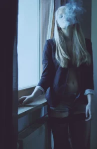 A young woman in a dark blazer exhaling smoke by a window, with the smoke partially obscuring her face, creating a moody and atmospheric portrait.