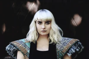 Intense gaze of a woman with platinum blonde hair and patterned shoulder pads, set against the dark background of a church door, presenting a strong fashion statement.