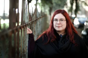 Confident woman with red hair and glasses standing by an old rusted fence, with soft-focus background highlighting her calm expression.