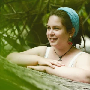 A smiling young woman with a headband and cherry earrings rests her arms on a log, surrounded by green foliage, radiating happiness and relaxation.