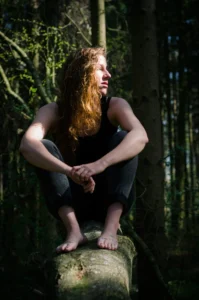 A woman with flowing hair sits thoughtfully atop a fallen tree in a sunlit forest, embodying a connection with nature.