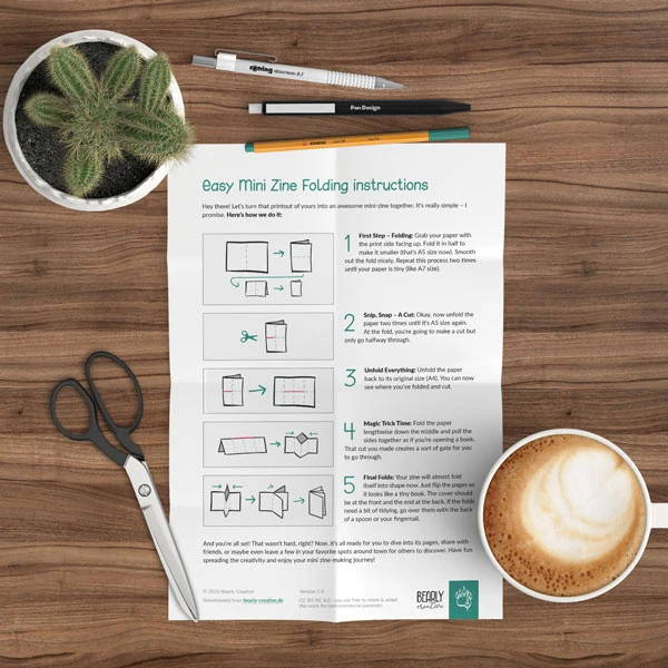 The image shows a set of visual instructions for folding a mini zine, laid out on a wooden surface. Surrounding the paper with the instructions are a pair of scissors, two pens, a mechanical pencil, a small potted cactus, and a cup of coffee with foam on top, suggesting a creative, DIY workspace. The instructions themselves include diagrams and text for each step, and the overall theme suggests a craft or hobbyist environment.
