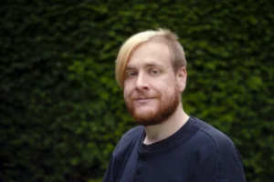 A smiling man with blonde hair and a beard, wearing a navy blue top, standing in front of a lush green hedge, in a casual outdoor portrait.