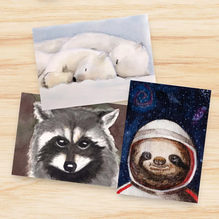 Photo of three artist trading cards on a wooden table. One of the cards shows two cuddling polar bears, one shows a raccoon and the third shows a sloth in a space helmet.