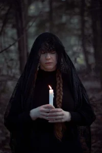 A solemn woman in a black lace veil holds a lit candle in a dark forest, her face partly obscured, creating a mood of introspection and mystique.