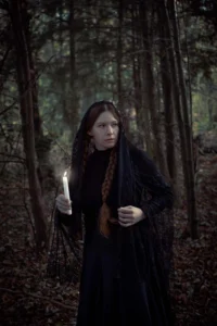 A woman draped in a black lace veil holds a candle in a dimly lit forest, her expression contemplative and serene amidst the natural surroundings.