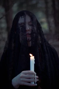 Woman shrouded in a black lace veil holding a lit candle in a dimly lit forest, creating a mysterious and contemplative mood.