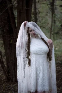 A young woman in a vintage white lace dress and veil, gently holding the veil in a forest setting, evoking a serene and ethereal atmosphere.