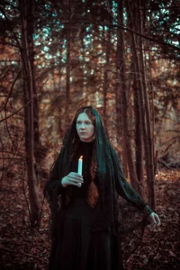 Mysterious woman in a dark cloak holding a lit candle in an autumnal forest, conveying a mood of enchantment and folklore.