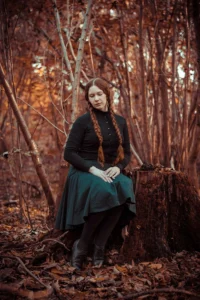 Photograph of a woman with red hair sitting on a tree stump in the forest. The colors are alienated - most of the picture is red, only her dark skirt has blue accents. She looks like she's daydreaming, her eyes are closed.