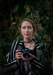 Confident female photographer holding a DSLR camera, framed by green foliage, with a focused and artistic expression.