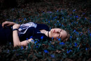 A young woman with fair hair lying amongst a field of vibrant blue flowers, her gaze upwards, blending with the serene twilight hues.