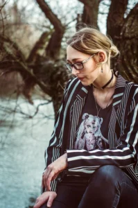 A thoughtful woman in stylish glasses and a striped shirt with a graphic print sits by a tree near a river, her expression serene and contemplative.