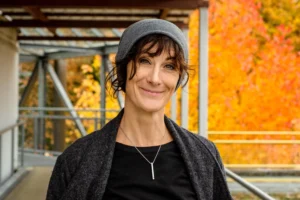 A smiling woman with a grey hat and cozy sweater stands outside, with autumn leaves providing a warm, golden backdrop.