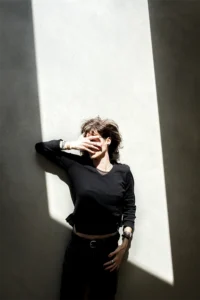 Top view of a woman dressed in black laying on a concrete floor. Her face is partially shaded by her hand from a streak of sunlight, creating a dramatic and artistic effect.