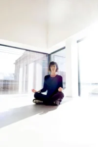 Woman meditating in a sunlit modern room with large windows, sitting cross-legged on the white floor, embodying calmness and mindfulness.