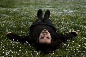 Man with dark hair wearing all black lying in a field of daisies, with his eyes closed and arms spread out, a peaceful expression on his face.